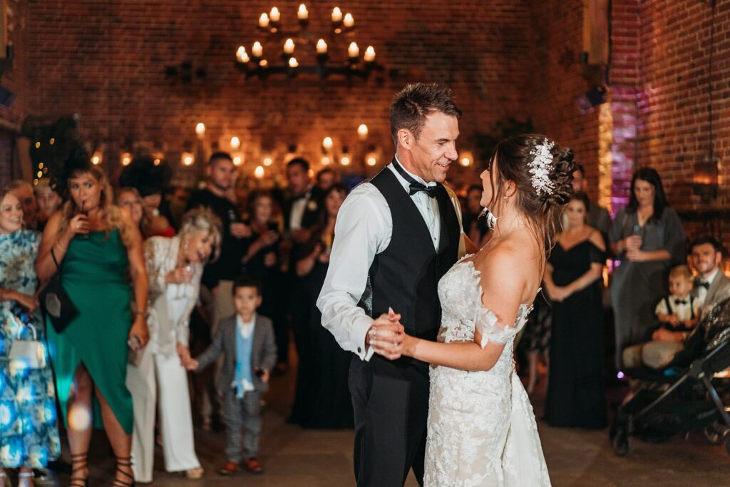 A bride and groom share their first dance in the warmly lit Hazel Gap Barn, smiling at each other as guests watch and take photos. Chandeliers and brick walls create a cozy, celebratory atmosphere.