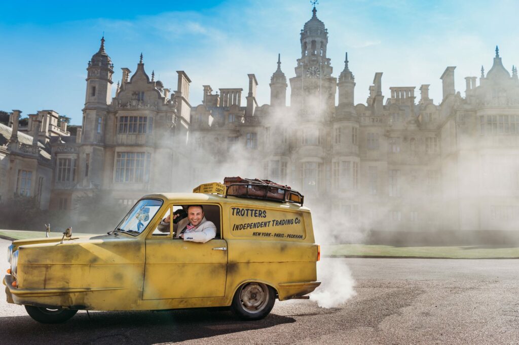 Groom talking on phone inside the Robin Reliant from Only fools and horses with Harlaxton Manor in the background