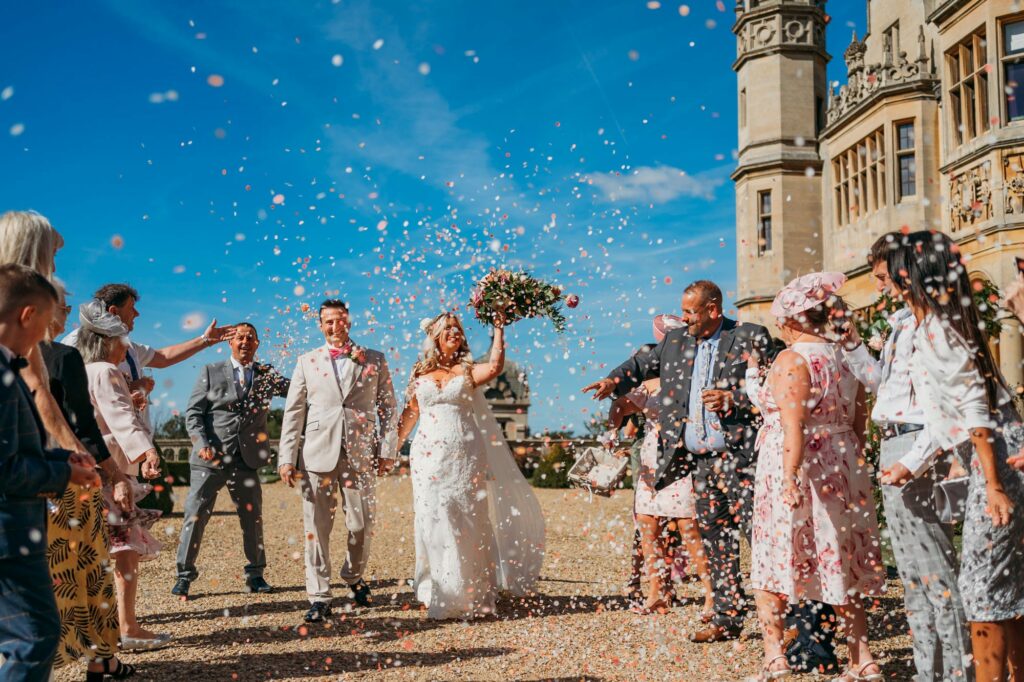 confetti being thrown at Harlaxton Manor as bride cheers with bouquet in the air