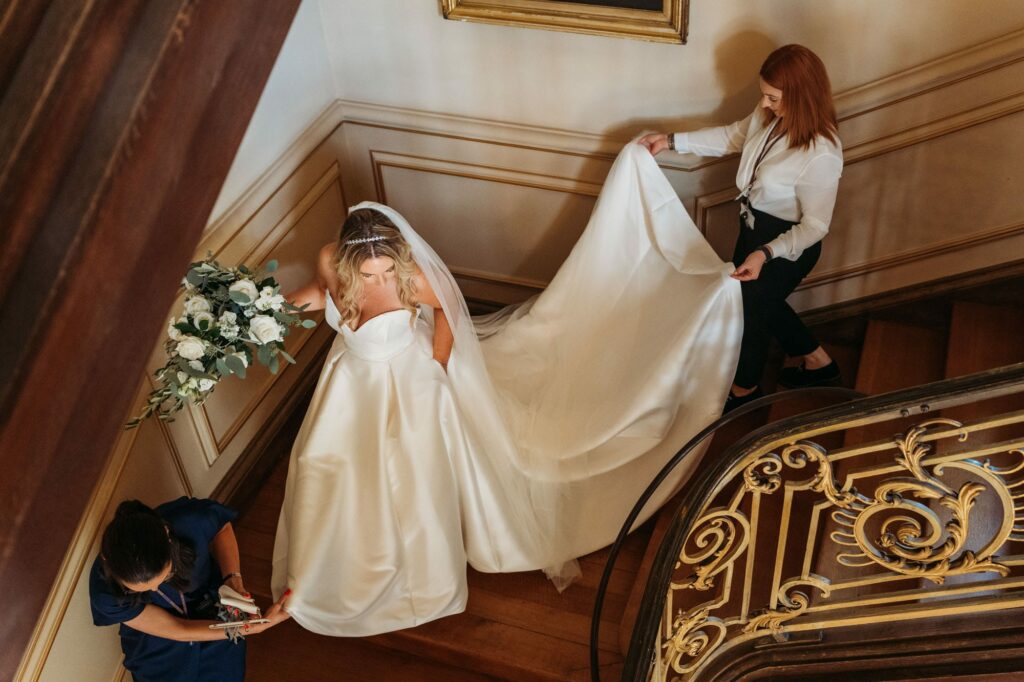 bride walking down the stairs at Harlaxton Manor to the wedding ceremony as staff help her with her wedding dress