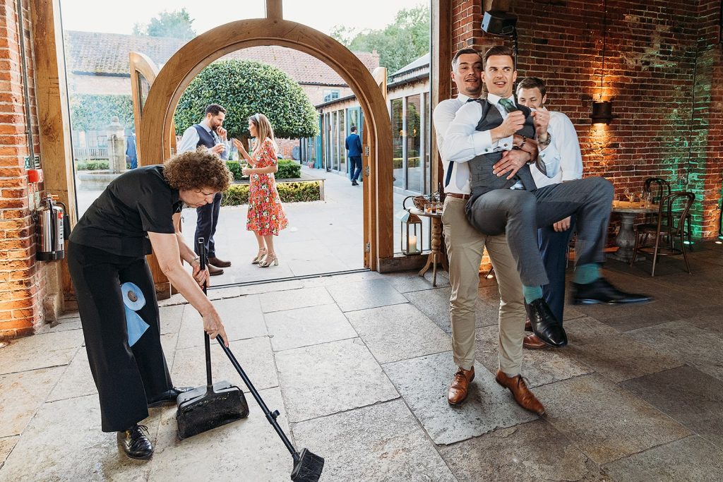 A person sweeps the floor indoors while two men, one lifting the other in his arms, pose playfully for the camera. In the background, two people talk outside in a garden area. The scene is lively and casual.