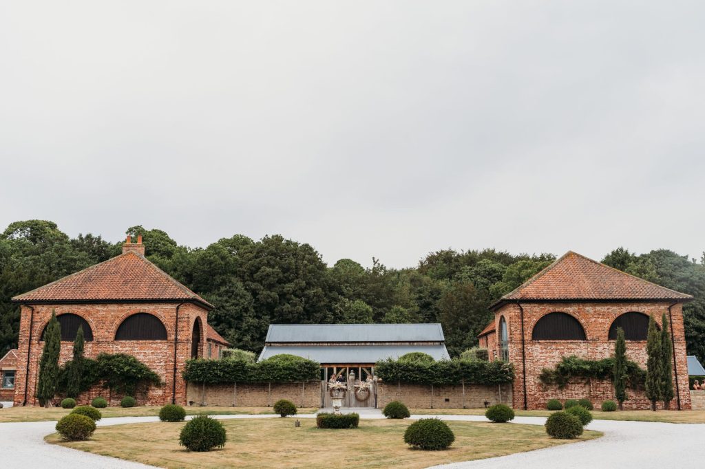 Two symmetrical watchtowers with arched windows flank a central wooden gate at Hazel Gap Barn, surrounded by trimmed bushes and a gravel path. Trees and greenery form the background under an overcast sky.