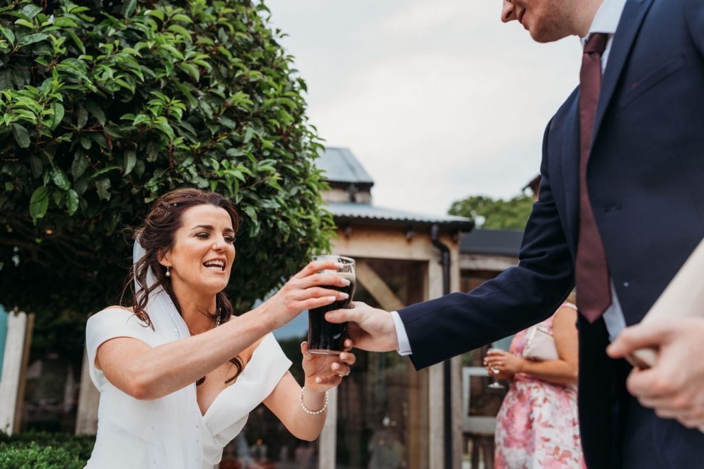 A bride in a white dress reaches to take a pint of dark beer from a man in a suit during an outdoor wedding celebration, with greenery and guests in the background.