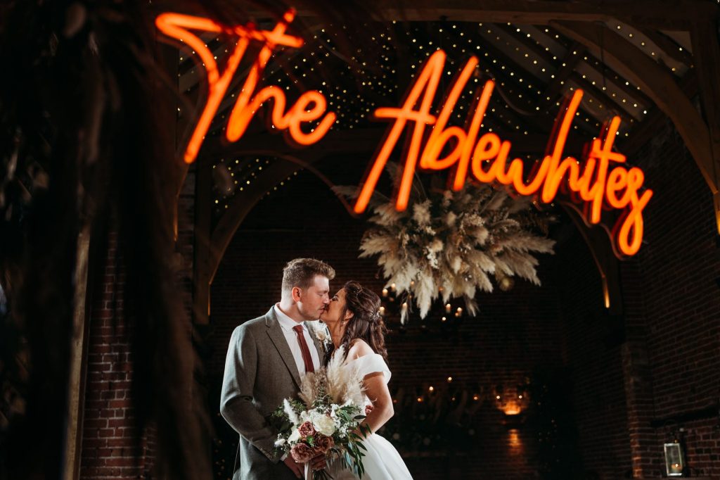 A bride and groom stand kiss under neon lights with their surname glowing saying The Ablewhites above them.