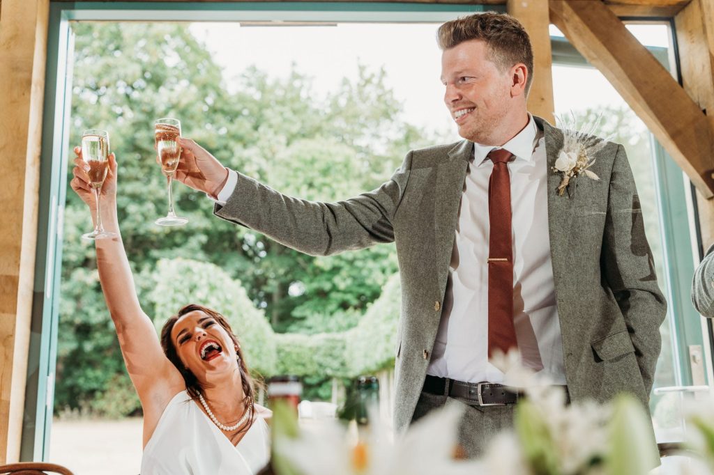 A cheering bride and groom raise champagne glasses in a celebratory toast, smiling enthusiastically inside Hazel Gap beside large windows and the garden visible outside.