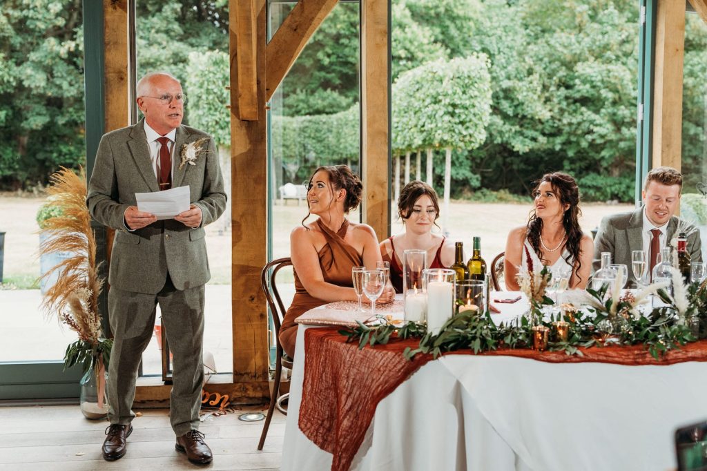 Father of Bride gives a speech at a wedding breakfast in the Oak Room at Hazel Gap Barn beside a decorated table, while three women and one man seated at the table smile and listen. Large windows and greenery are visible in the background.