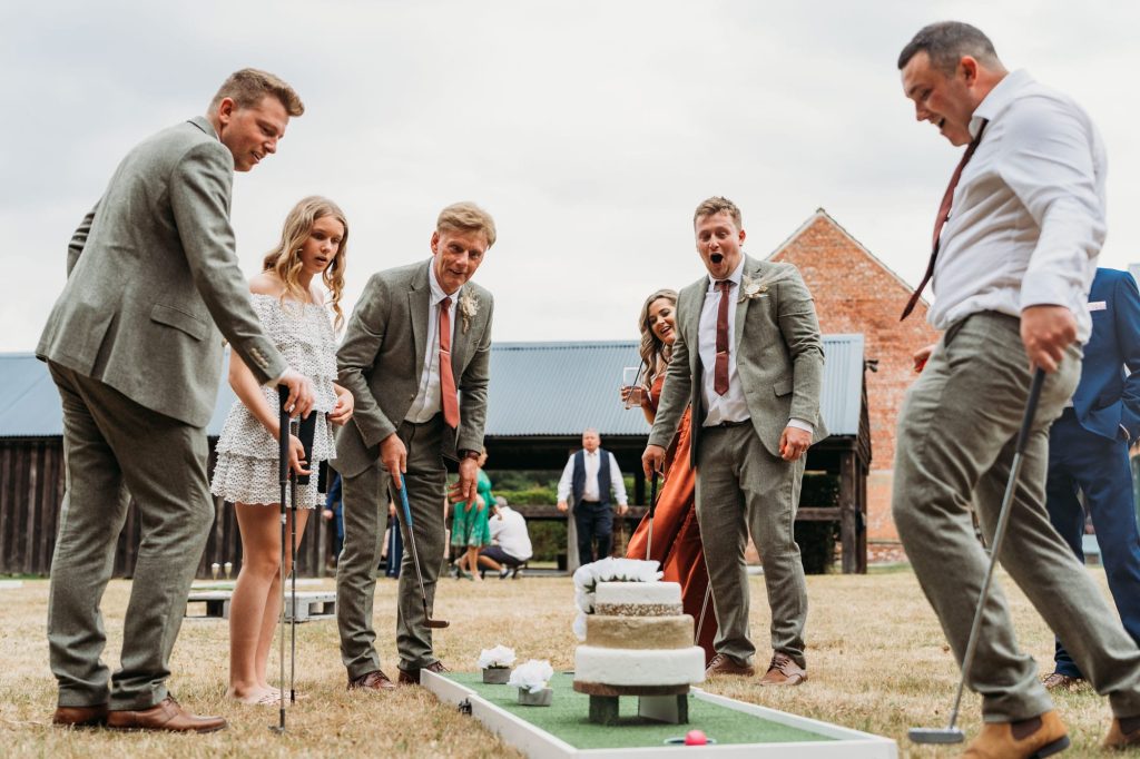 A group of people dressed in semi-formal attire excitedly play mini-golf outdoors on grass, reacting with enthusiasm around a small putting green with a wedding cake obstacle.