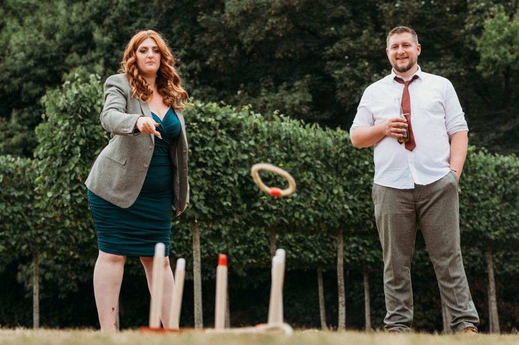 A woman in a green dress and gray blazer tosses a ring in an outdoor lawn game, while a man in a white shirt and loosened tie stands beside her, watching and holding a drink. Trees and bushes are in the background.