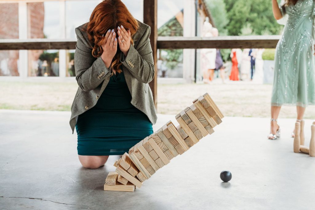 A woman kneels on the ground covering her face with her hands as a large wooden Jenga tower collapses in front of her. A black ball rests nearby, and people are visible in the background.