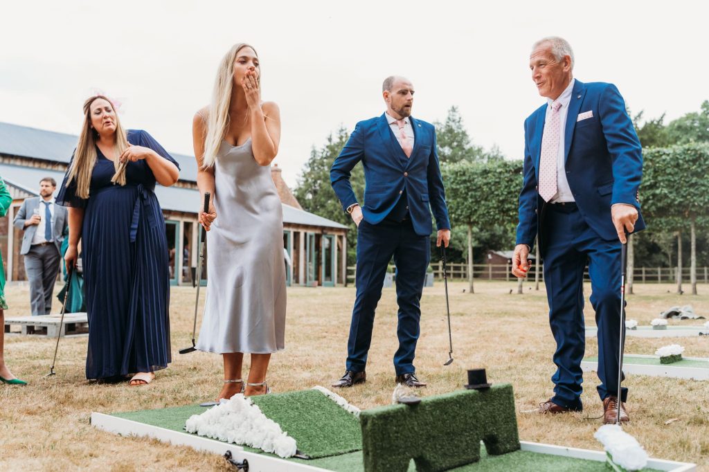 A group of people dressed in formal attire play mini-golf outdoors. A woman in a silver dress blows a kiss, while others watch and hold golf clubs. Trees and a building are visible in the background.