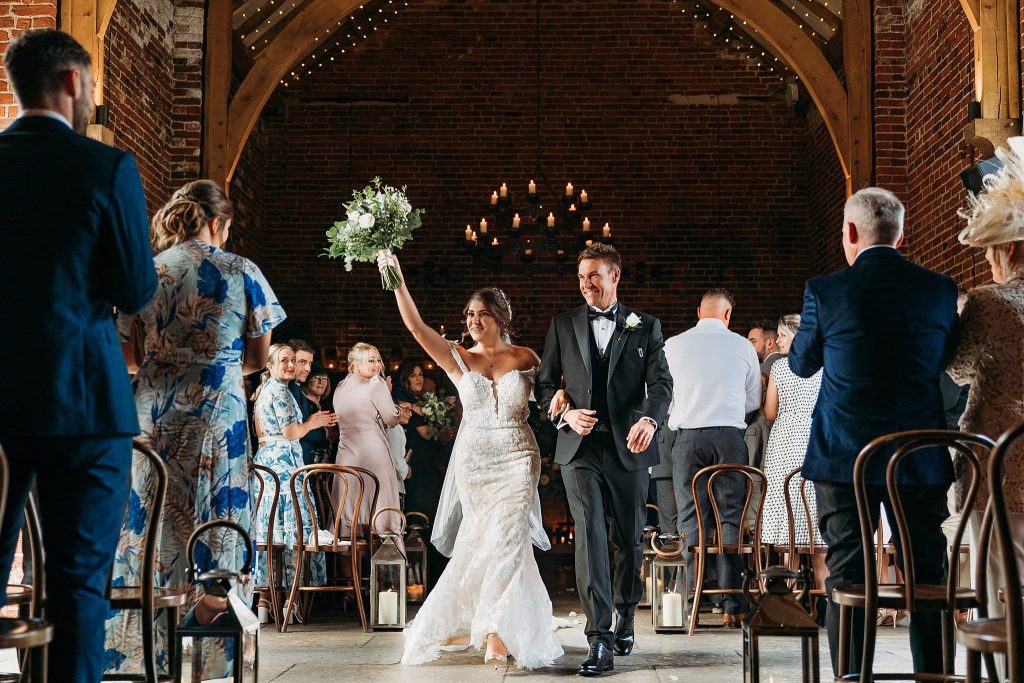 A bride in a white gown holds a bouquet up triumphantly while walking down the aisle with her groom in a black tie tuxedo; guests stand and smile inside Hazel Gap Barn’s rustic, warmly lit venue.