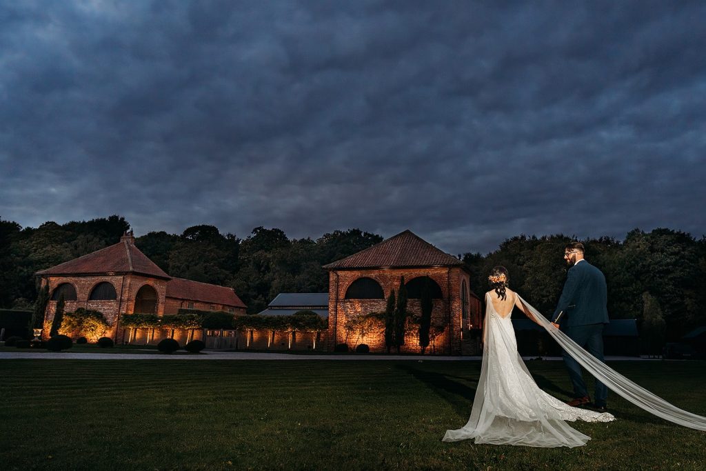 A bride and groom walk back to Hazel Gap Barn at dusk, holding hands, with a dramatic dark blue clouds gather overhead