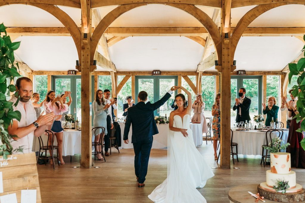 A bride and groom dance in the centre of the Oak room at Hazel Gap Barn as guests stand, clap, and smile. Tables are decorated for a wedding, while sunlight streams through large windows in the background.