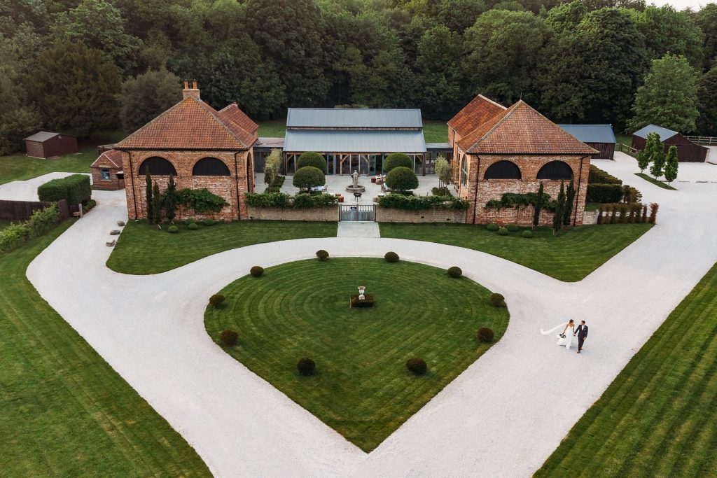 A bride and groom walk along a curved gravel driveway in Aerial drone photo of bride and groom walking around the path at Hazel Gap Barn with the venue clearly visible in the top of the frame