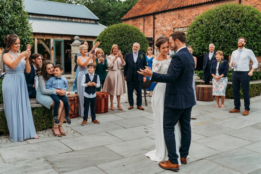 A newlywed couple dances outdoors on a stone patio, surrounded by smiling guests who are clapping and taking photos.