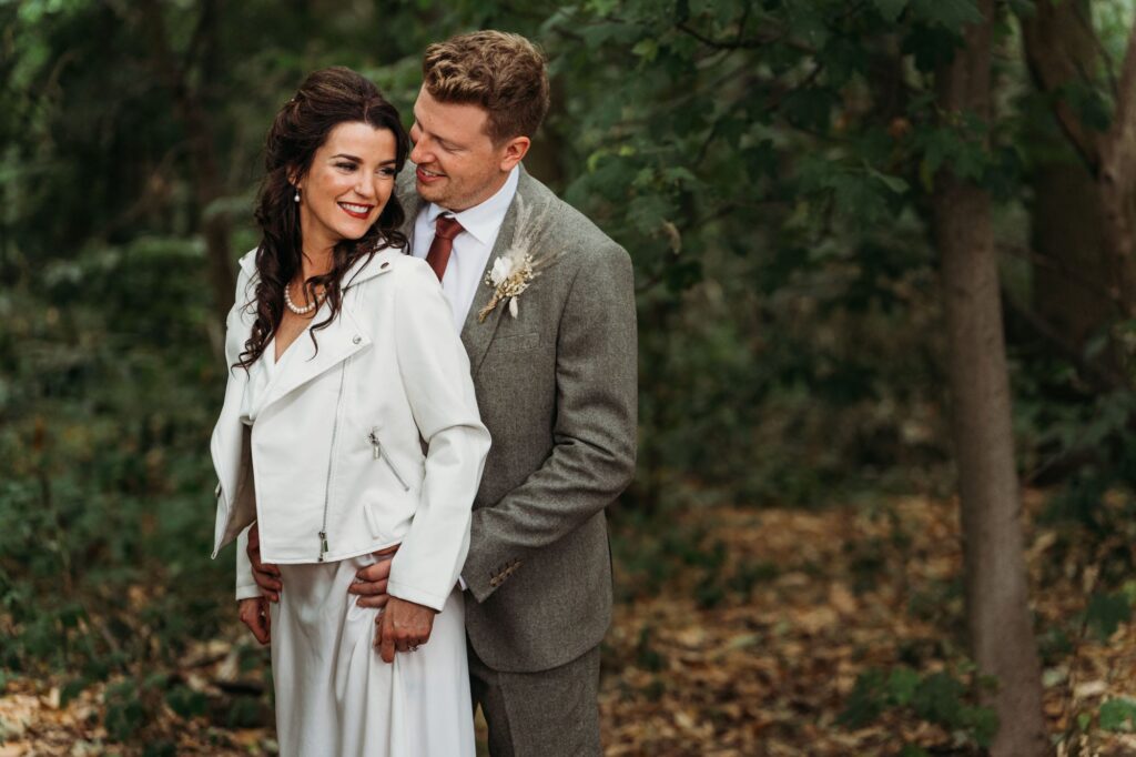 bride wearing a custom leather jacket for her wedding standing in the woods with groom hugging her