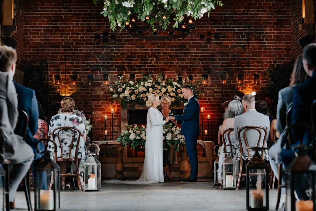 A bride and groom stand facing each other, exchanging rings during a wedding ceremony indoors at Hazel Gap Barn, with guests seated, floral arrangements, candles, and greenery decorating the background.