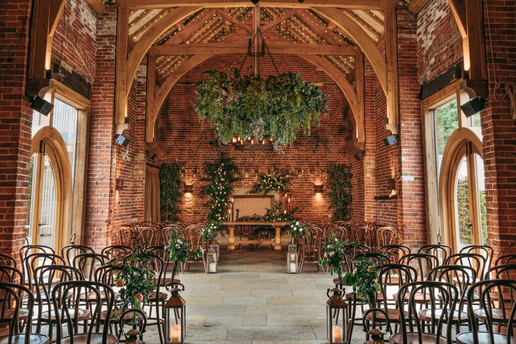 Rustic wedding ceremony setup at Hazel Gap Barn with wooden beams, rows of chairs, greenery and floral decorations, lanterns lining the aisle, and a large hanging arrangement of foliage overhead.