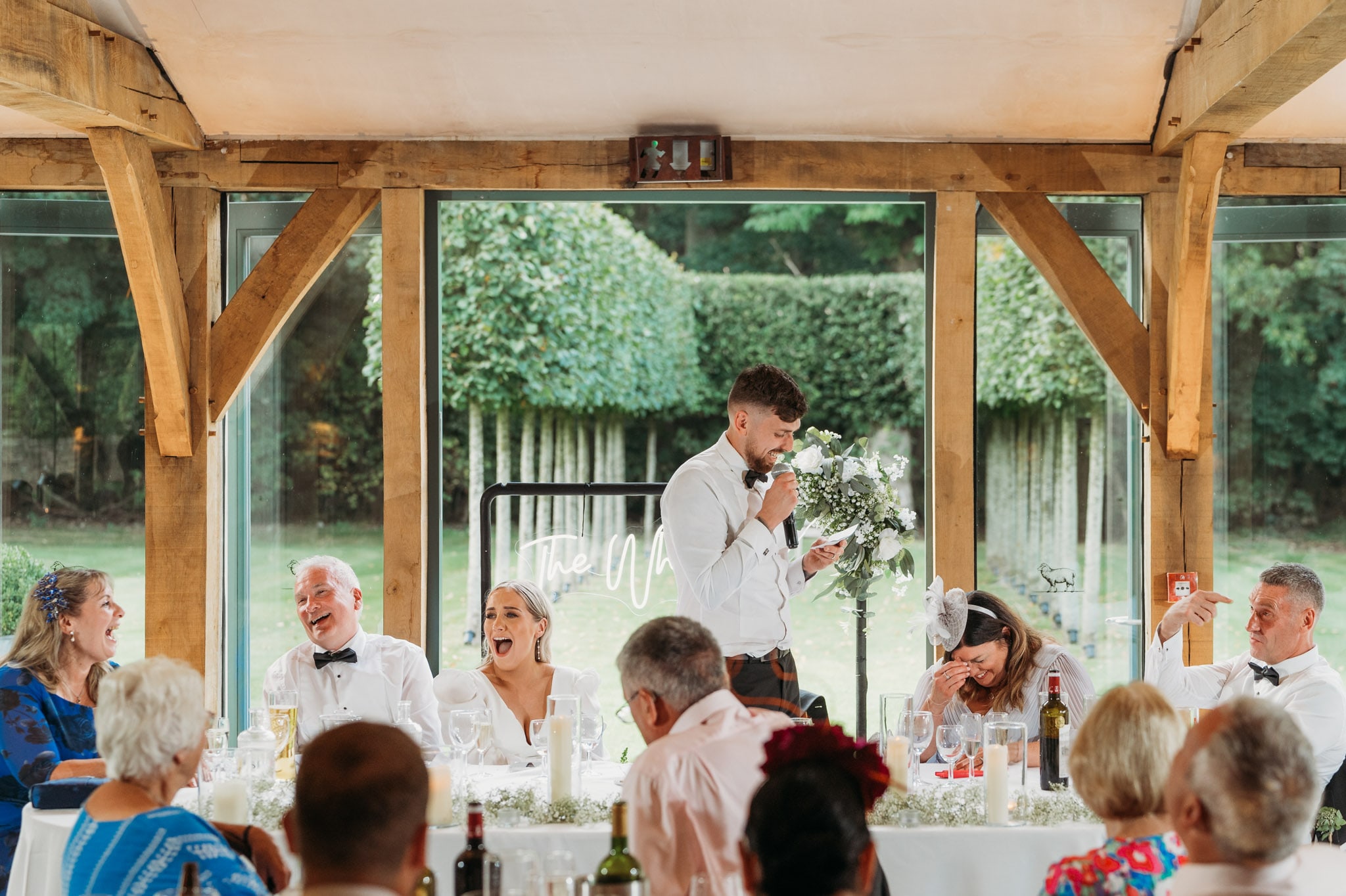 Groom giving a speech at a Hazel Gap Barn wedding reception, making guests laugh. People are seated at a long table, smiling and reacting with joy, with greenery visible through large windows behind them.