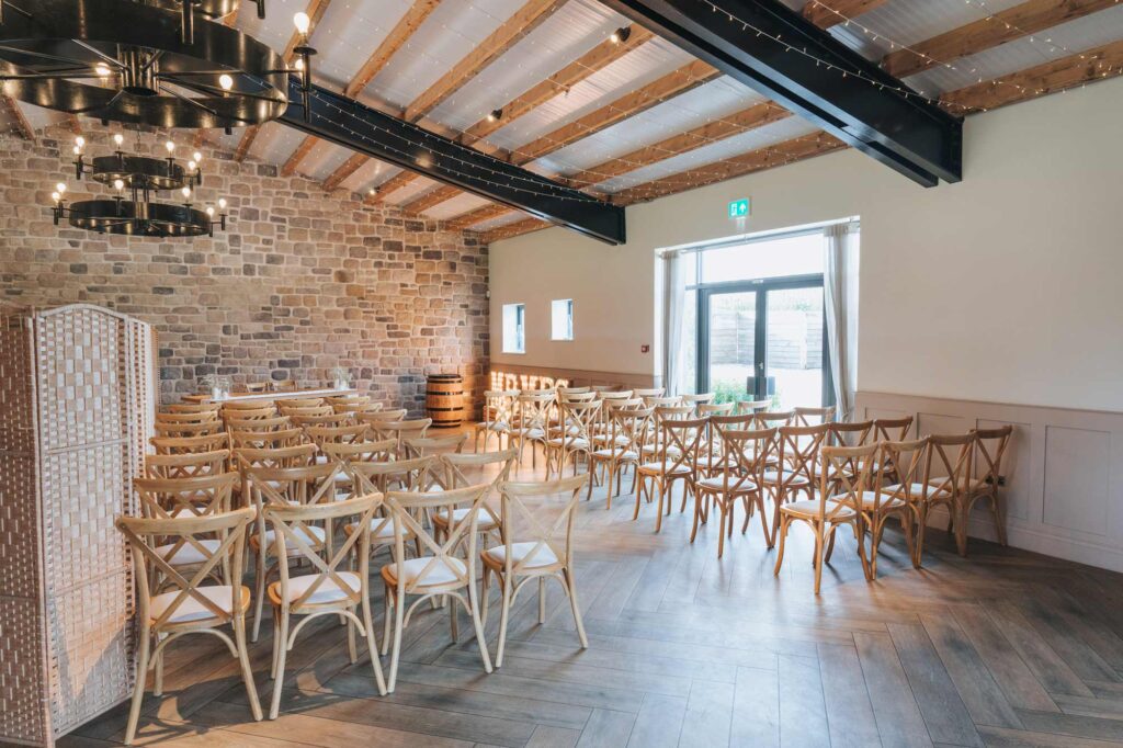 Elegant event space with rows of wooden cross-back chairs facing a stage partition, exposed beam ceiling with industrial lighting, stone walls, and a large window providing natural light.