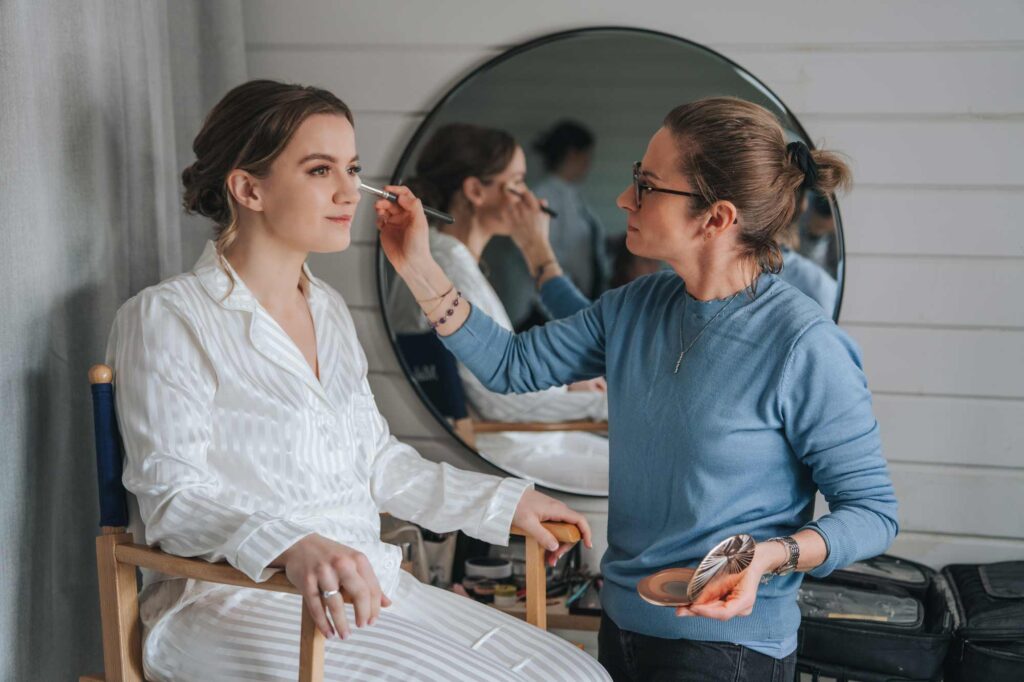 A makeup artist is applying makeup to a seated woman in a white striped pajama set; they are both reflected in a large round mirror in a well-lit room.