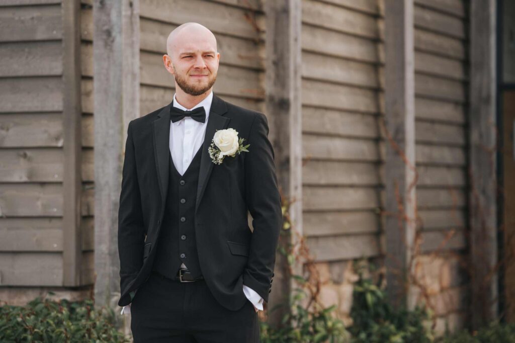 A groom wearing a black tuxedo with a bow tie and a boutonniere, standing in front of a Stretton Manor Barn with a subtle smile.