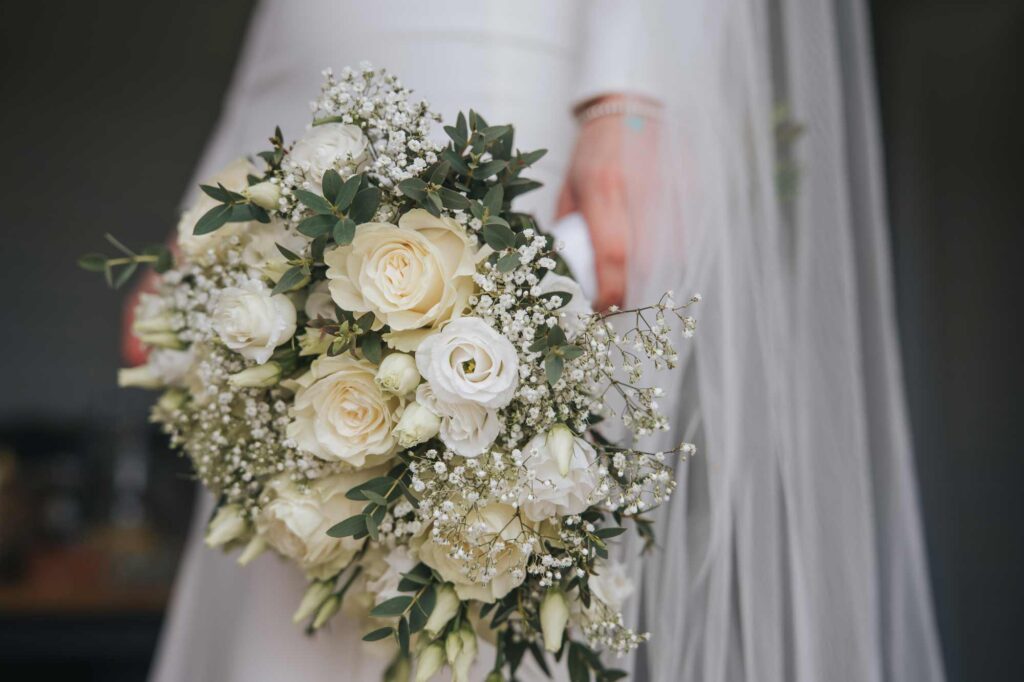 A close-up photo of a bride holding a bouquet of white roses and baby's breath, with a glimpse of a white wedding dress and veil in the background.