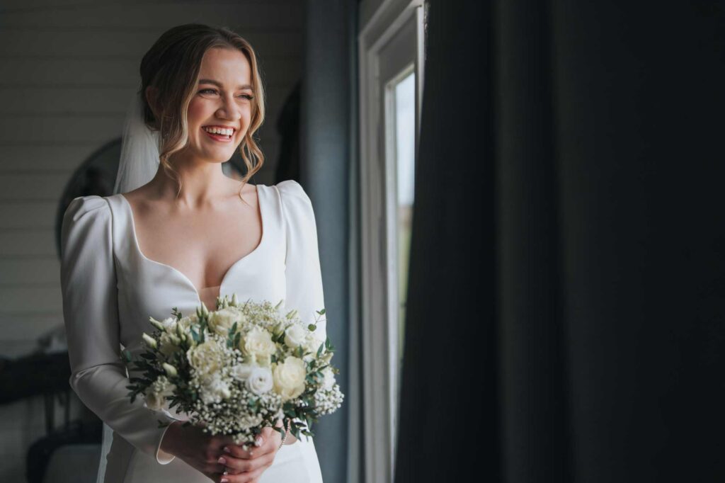 A smiling bride in her wedding dress holding her a bouquet of flowers, standing by a window.