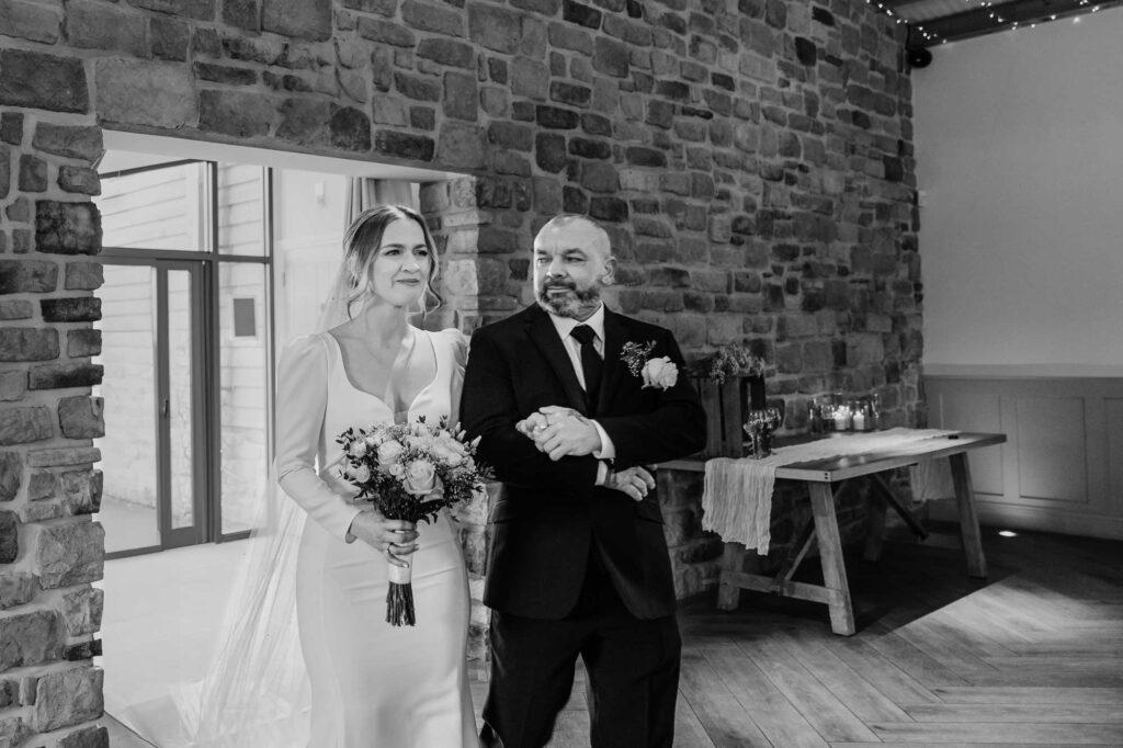 A black and white photo of a bride holding a bouquet walking arm in arm with father of bride wearing a suit, both are standing inside a room with rustic stone walls and wooden floor.
