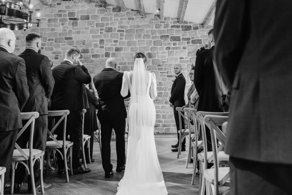 A black and white photo of a bride walking down the aisle while guests stand either side, viewed from behind the bride.