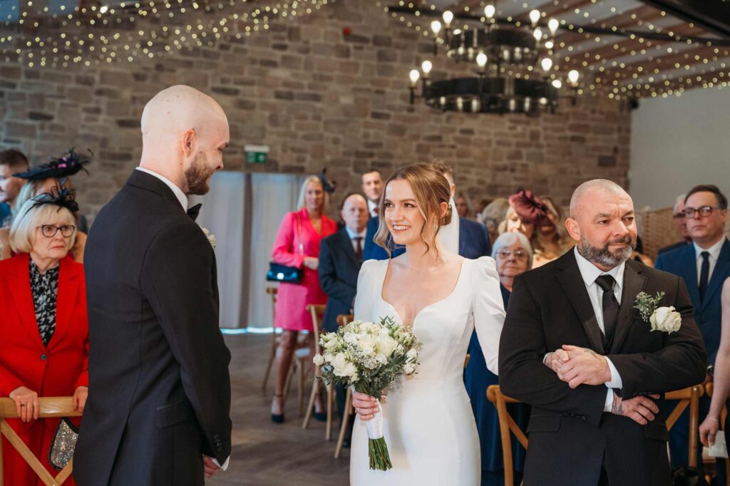 A wedding ceremony in a room with stone walls and hanging string lights. A bride in a white dress holding a bouquet is smiling at a groom in a black suit. An older man in a black suit appears emotional, holding his hands together in the foreground. Guests are seated on either side, observing the ceremony.