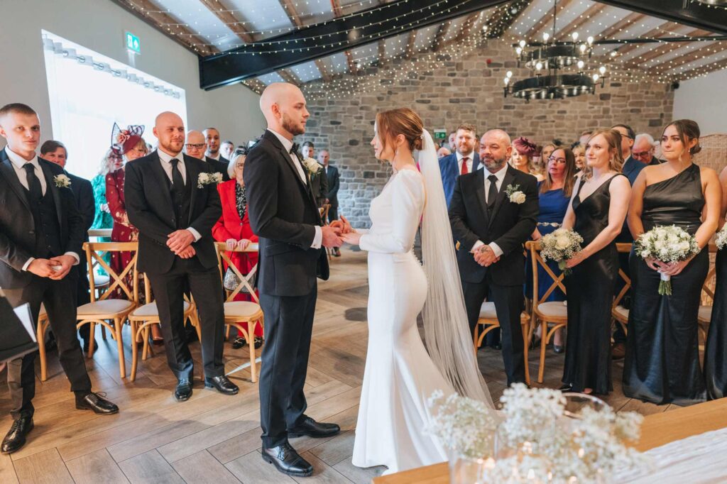 A bride and groom holding hands during their wedding ceremony, surrounded by guests and bridesmaids dressed in black, in a room decorated with string lights and stone walls.