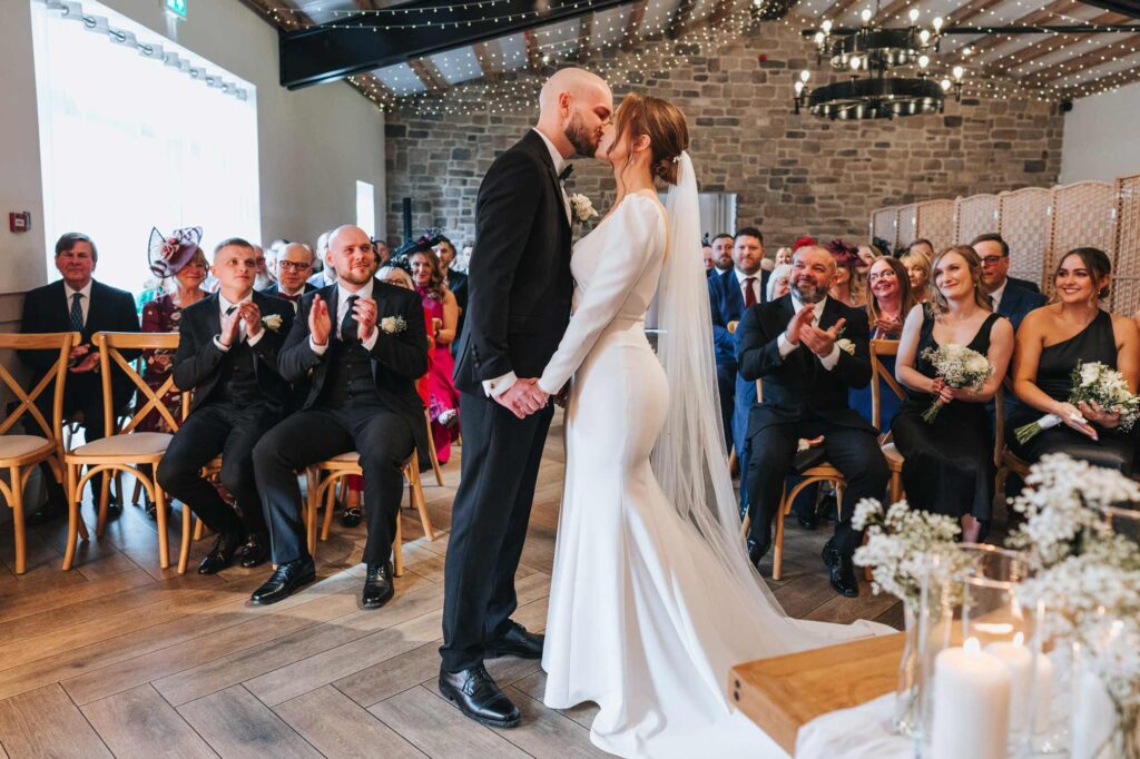 A bride and groom kissing in front of wedding guests who are clapping. The setting features a rustic interior with stone walls and string lights.