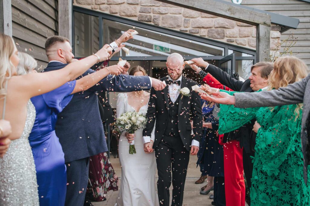 A bride and groom holding hands while walking through a shower of confetti thrown by wedding guests outside of Stretton Manor Barn