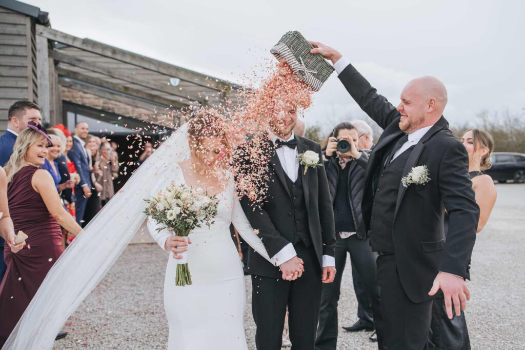 A bride and groom holding hands while smiling and walking, as confetti is joyously thrown over them, with guests smiling and capturing the moment in the background.