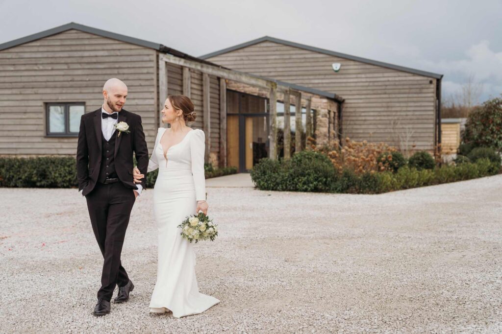 groom in black tuxedo walking with bride holding a bouquet with Stretton Manor Barn, a Derbyshire wedding venue behind them