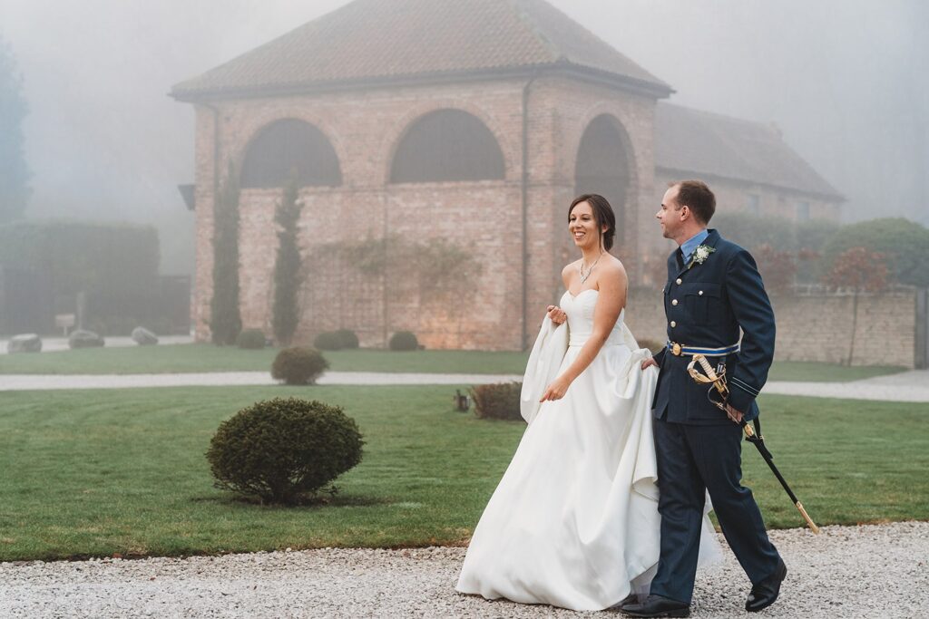 A bride in a white gown and a groom in a formal military uniform walk together outside on a gravel path, smiling, with a brick building and green lawn in the background.