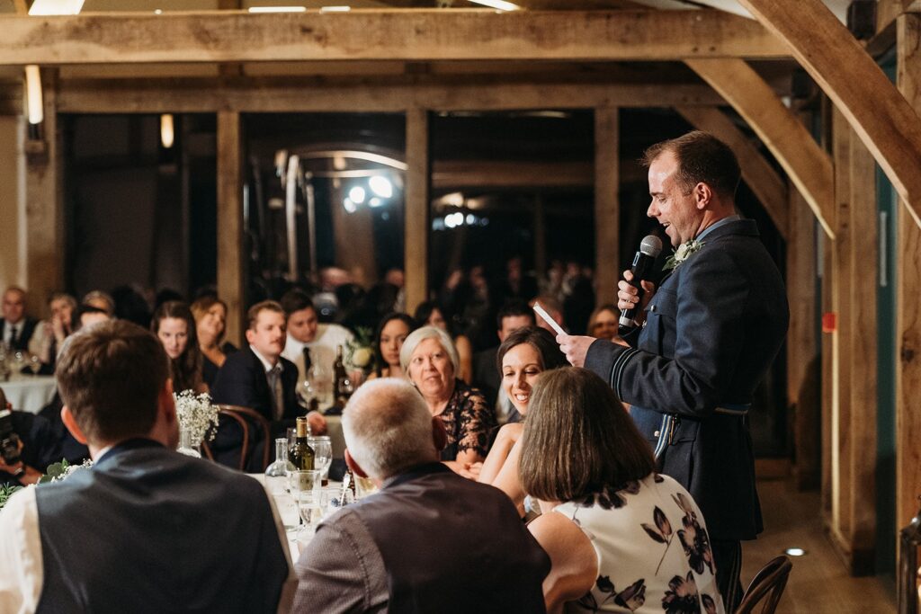 Groom in RAF dress uniform gives a speech in winter at Hazel Gap's Oak room. Outside is dark/night time