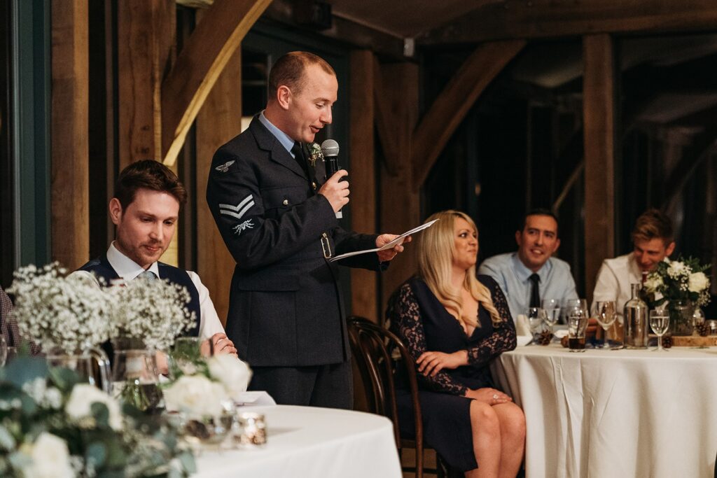 Groom in RAF dress uniform gives a speech in winter at Hazel Gap's Oak room. Outside is dark/night time