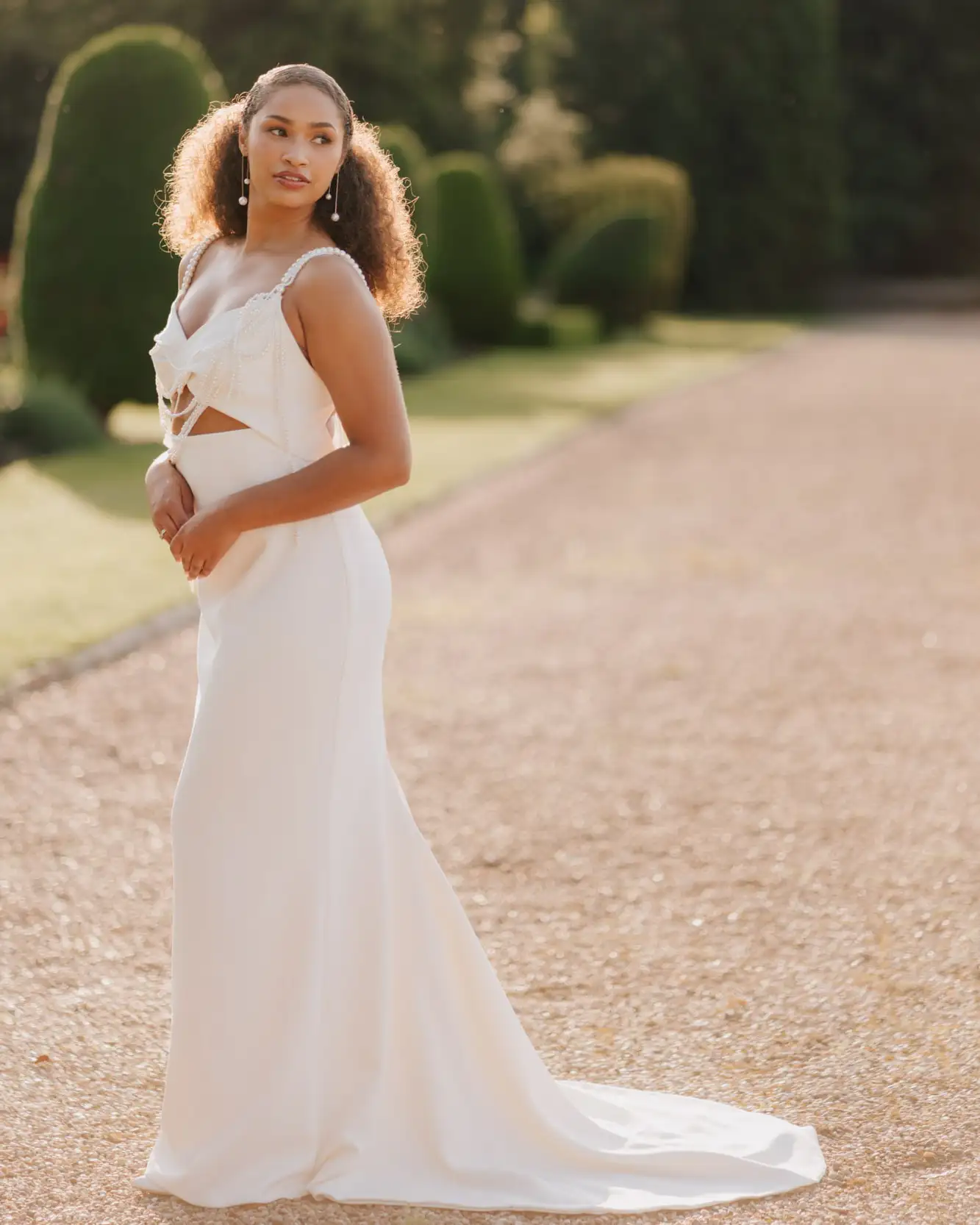 A bride in a white, floor-length, sleeveless bridal gown with lace details stands outdoors on a gravel path, looking to the side. Captured by a Nottingham Wedding Photographer, green hedges and sunlight create a soft, elegant background.