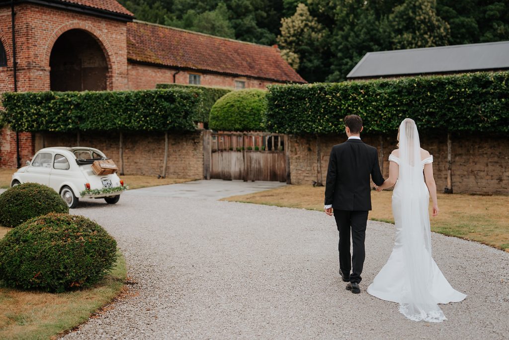A bride in a white dress and groom in a black suit walk hand-in-hand down a gravel path, with a vintage white car and Hazel Gap Barn in the background.