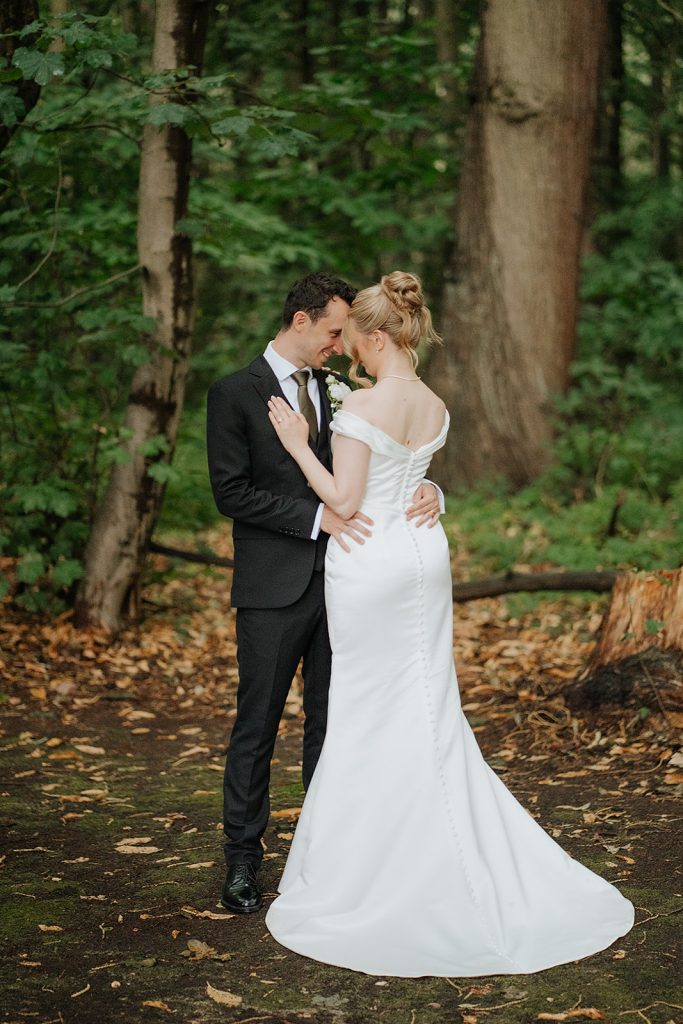 A bride in a white gown and a groom in a black suit embrace in a forest, touching foreheads and surrounded by green trees and fallen leaves.