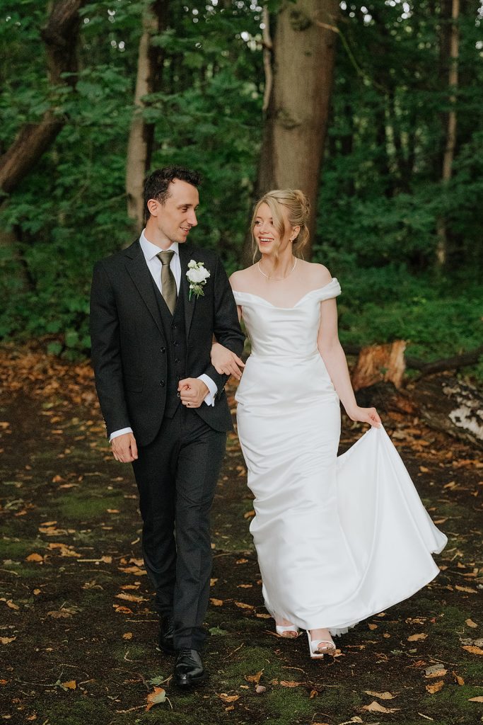 A bride in a white dress and a groom in a dark suit walk arm in arm through a wooded area, smiling at each other, with green trees and fallen leaves in the background.