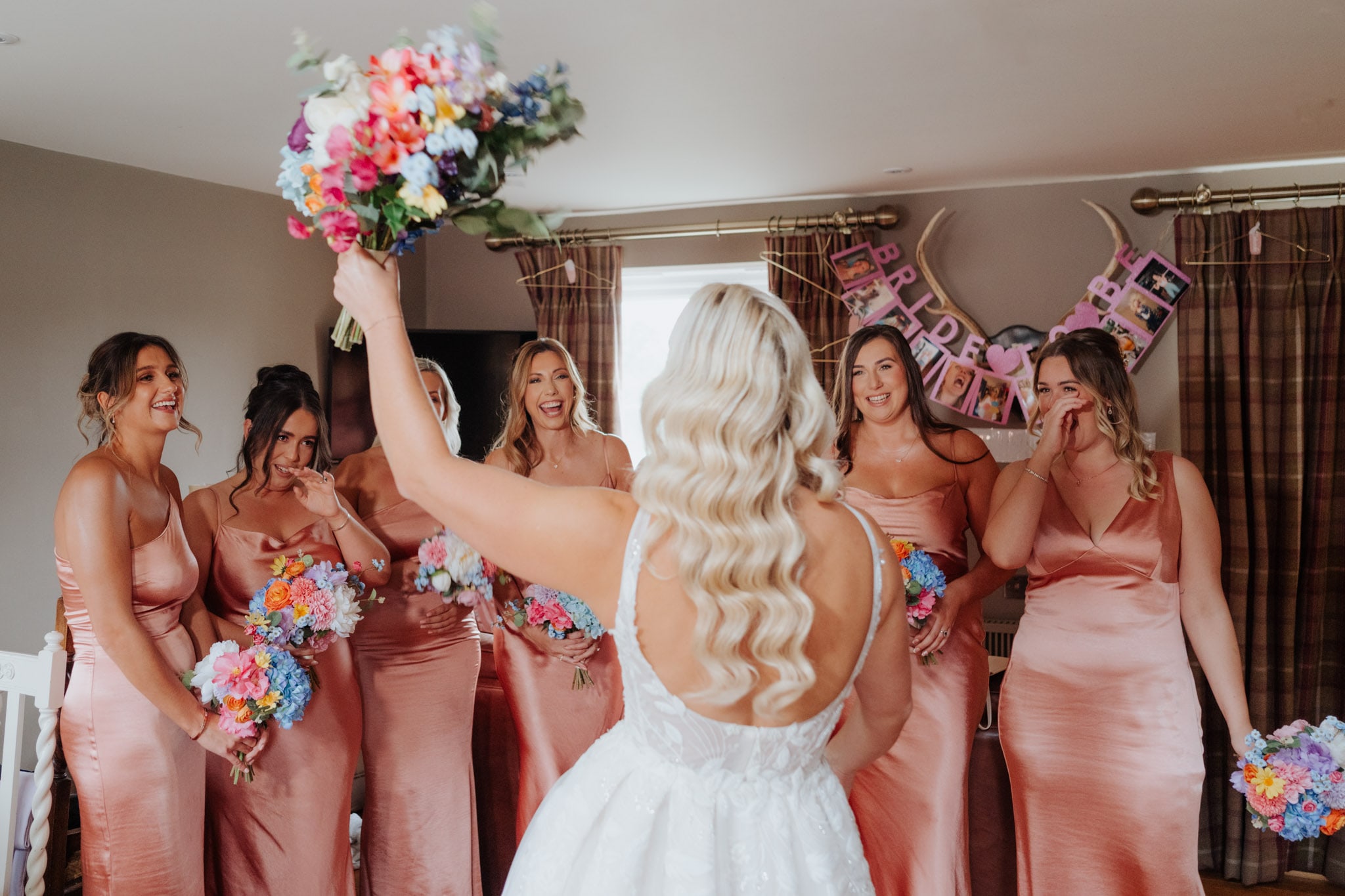 A bride in a white dress faces five smiling bridesmaids in matching blush dresses, holding colorful bouquets. The bride raises her bouquet as the bridesmaids react joyfully. A BRIDE TO BE banner hangs in the background.