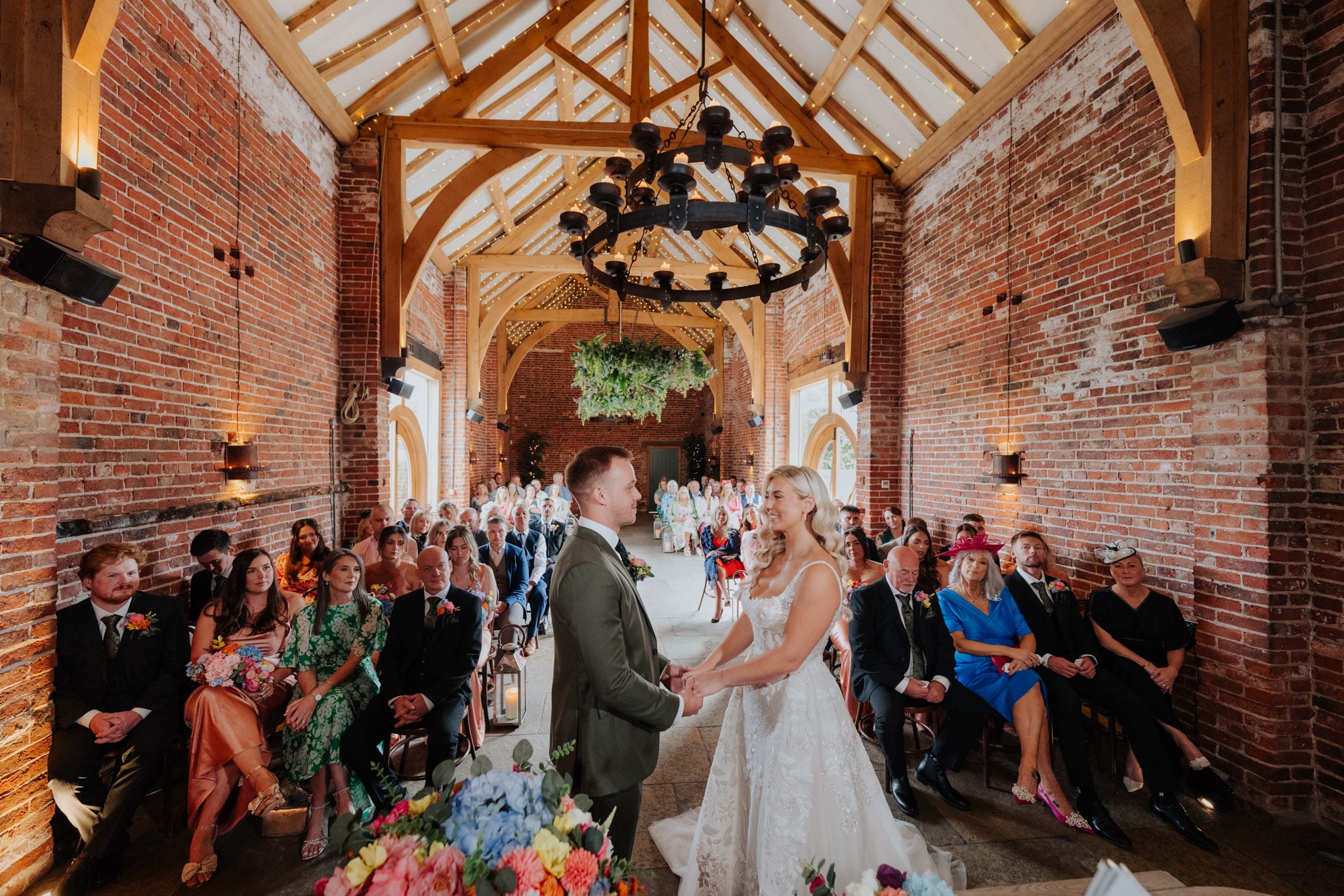 A bride and groom stand holding hands at the altar in Hazel Gap Barn surrounded by seated guests. The ceiling features exposed wooden beams and a central chandelier. Flowers decorate the scene.