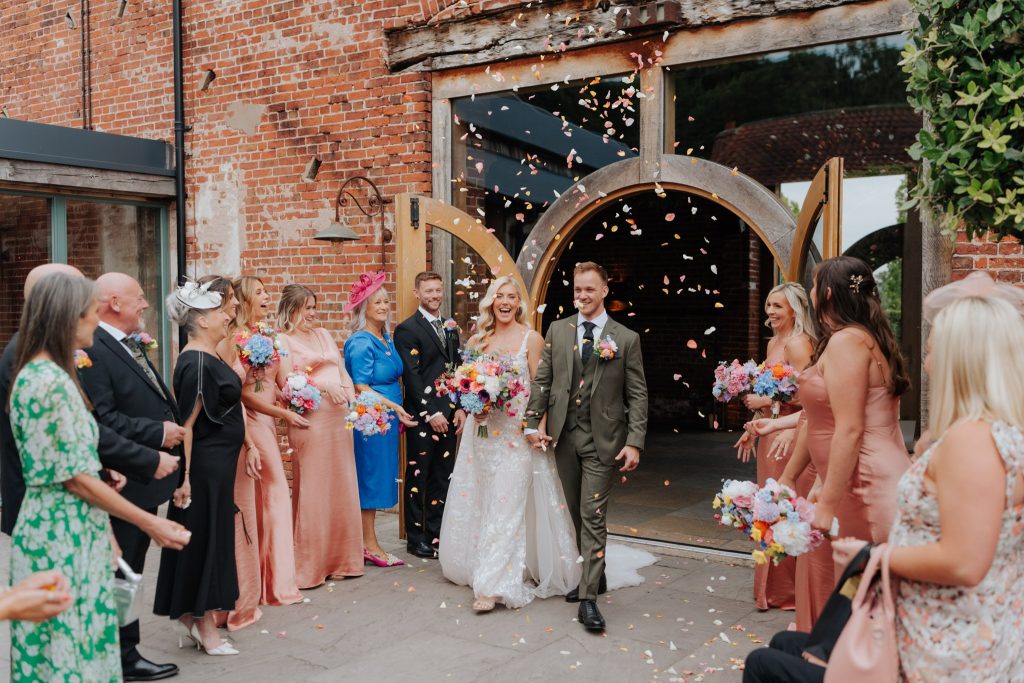 A bride and groom smile and walk hand-in-hand outside a rustic brick building as guests toss flower petals. Bridesmaids in pink dresses and guests in formal attire look on, celebrating the newlyweds.
