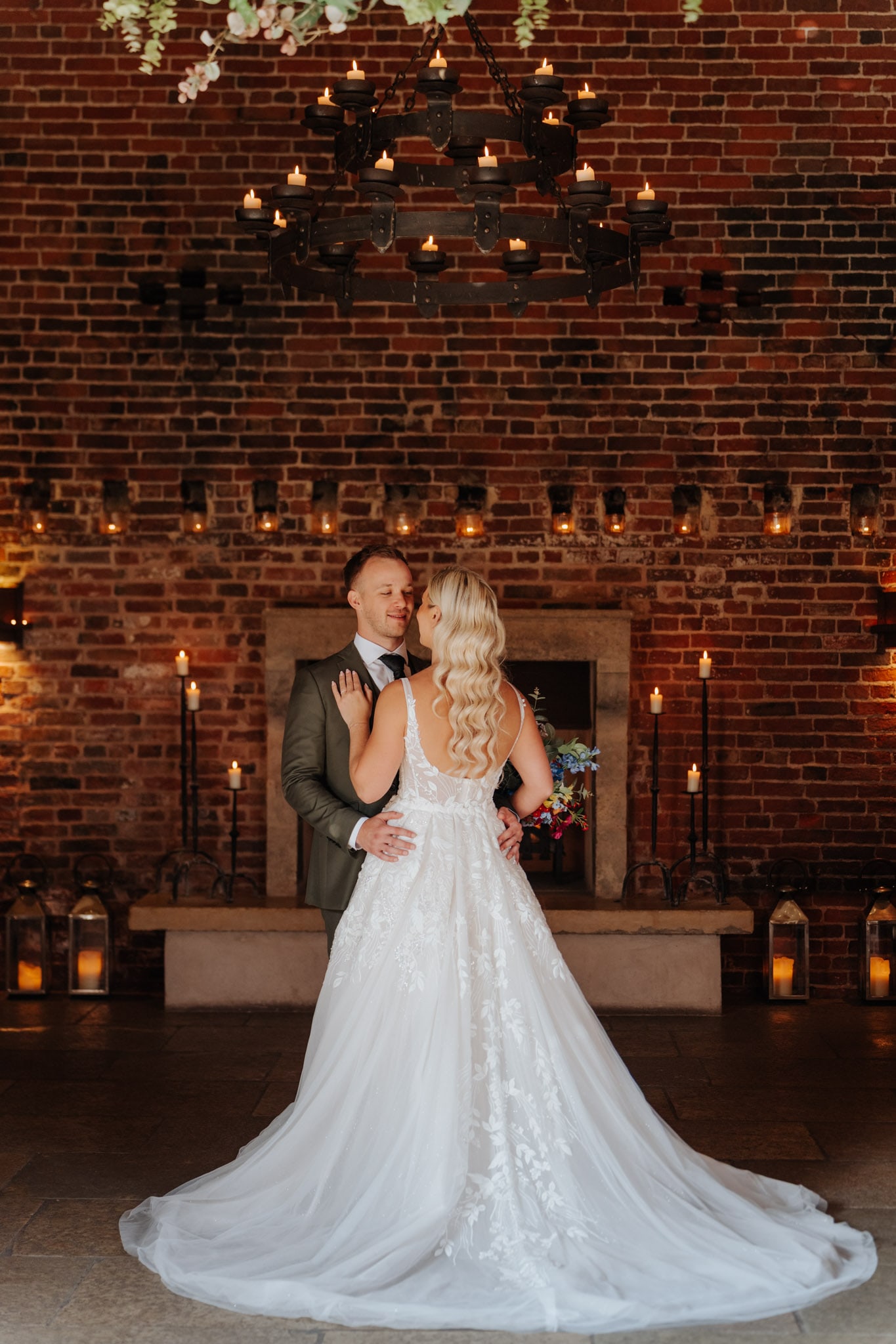 A bride in a white gown and a groom in a suit stand facing each other in front of a brick wall and fireplace, surrounded by candles and lanterns, under a large chandelier.