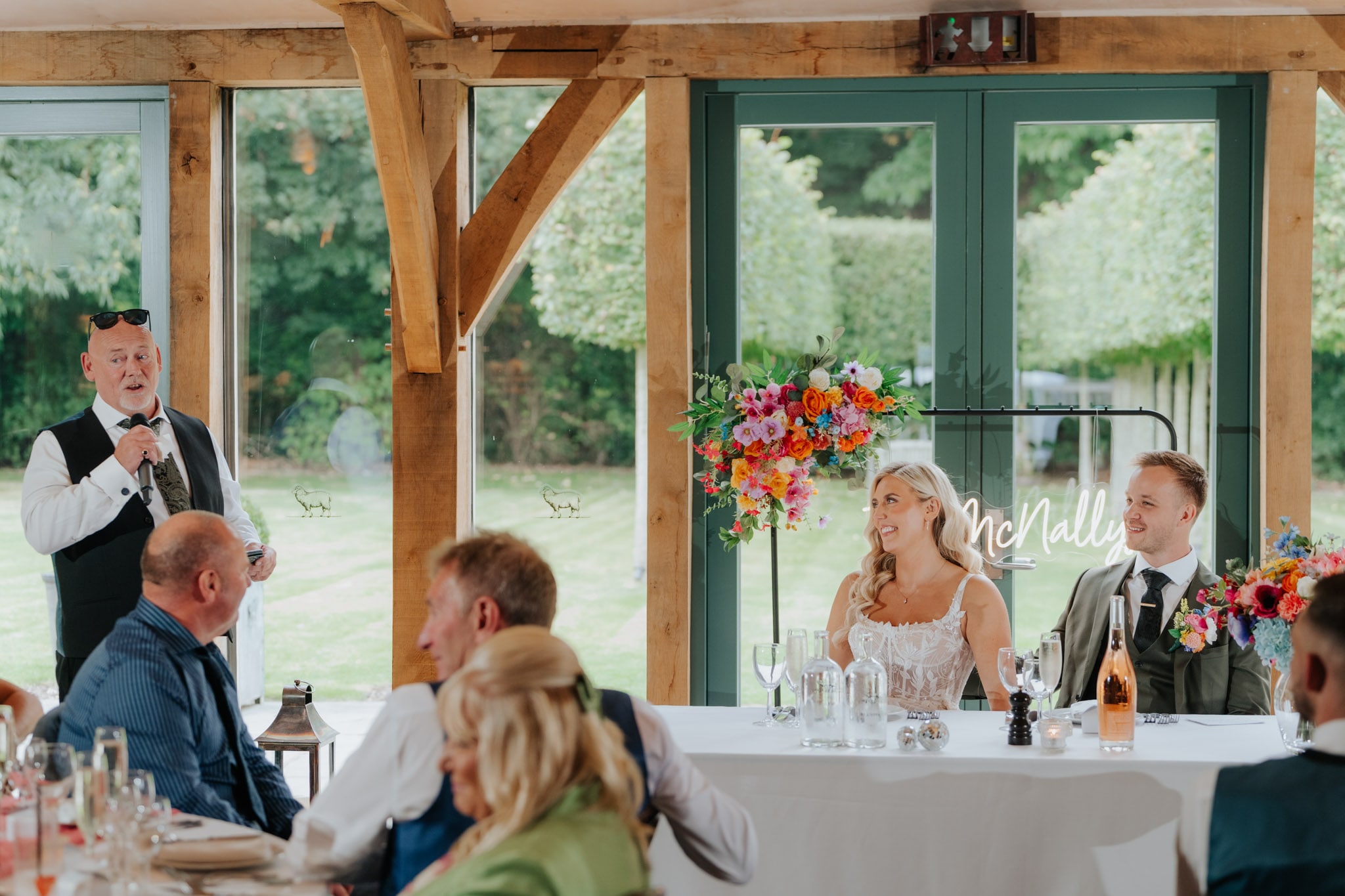 A man in a vest and sunglasses gives a speech with a microphone at a wedding reception, while the bride and groom sit smiling at a decorated table in a light-filled room with large windows and flowers.