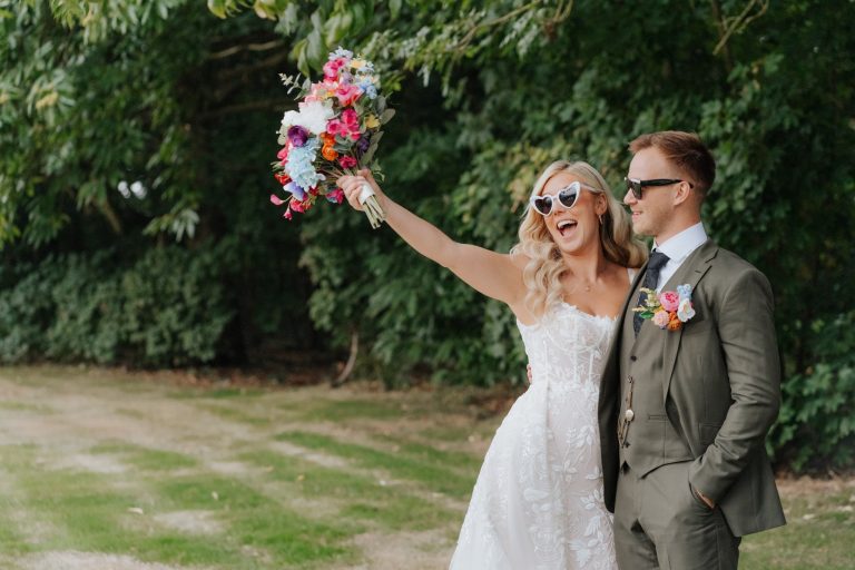 A bride in a white dress and heart-shaped sunglasses holds up a colorful bouquet, smiling next to a groom in a green suit and sunglasses, standing outdoors with greenery in the background.