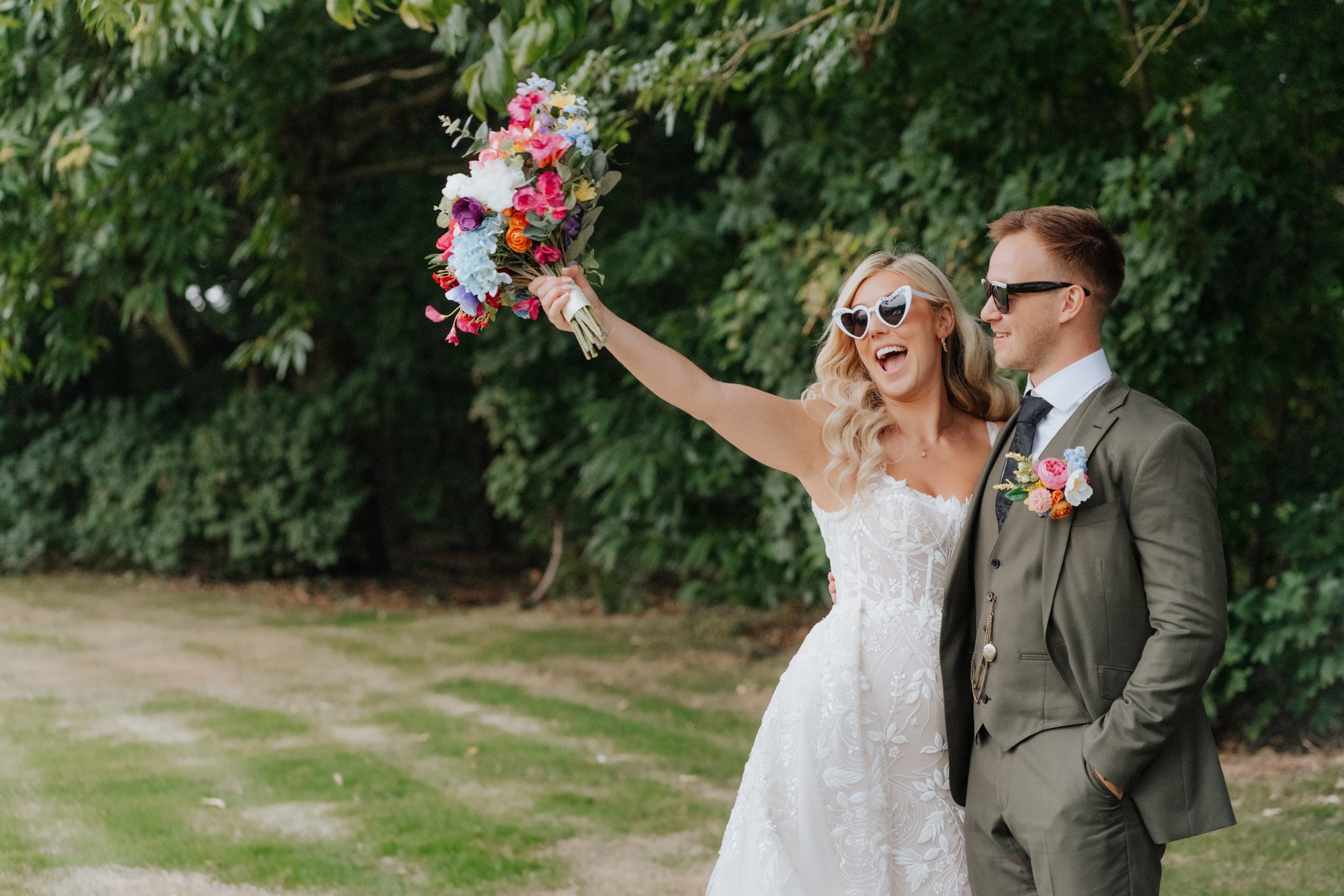 A bride in a white dress and heart-shaped sunglasses holds up a colorful bouquet, smiling next to a groom in a green suit and sunglasses, standing outdoors with greenery in the background.